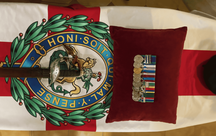 A military sword, medals on a red cushion, and a hat rest on a flag-draped coffin featuring the Order of the Garter emblem and Honi Soit Qui Mal Y Pense motto—paying tribute to a distinguished Military Veteran.